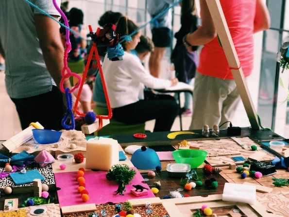 A table with arts and craft items scattered over it. Children in the background, slightly blurred.